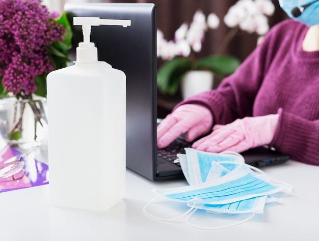 Foto alcohol ontsmettingsgel voor handhygiëne, covid-19 coronavirus coronabescherming, medische chirurgische blauwe gezichtsmaskers. vrouw met masker, handschoenen werken vanuit huis met behulp van laptop. medicijnen, gezondheidszorg