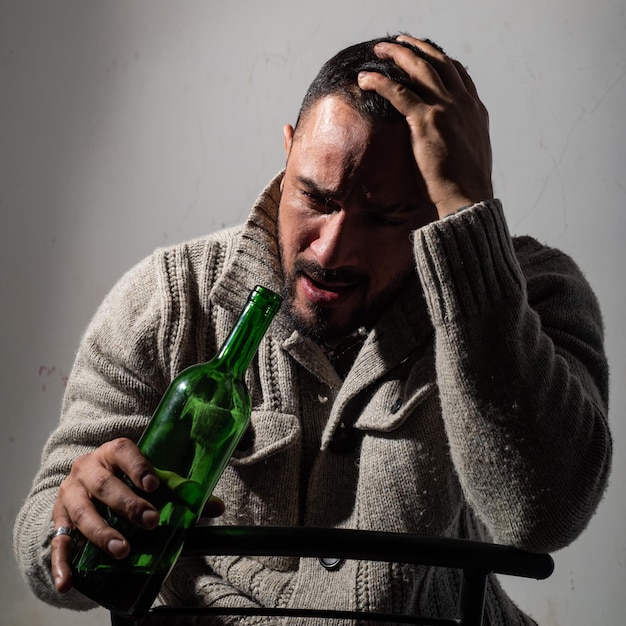 알코올 중독 알코올 중독과 사람들의 개념 강모가 있는 좌절된 남자가 울면서 머리를 잡고 손에 병을 들고 앉아 있습니다.