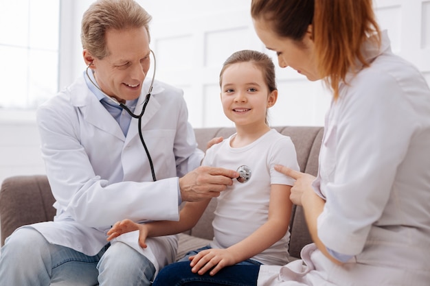 All attention is yours. Prominent gentle charming doctor running a general medical checkup examining girls lungs while his colleague comforting her