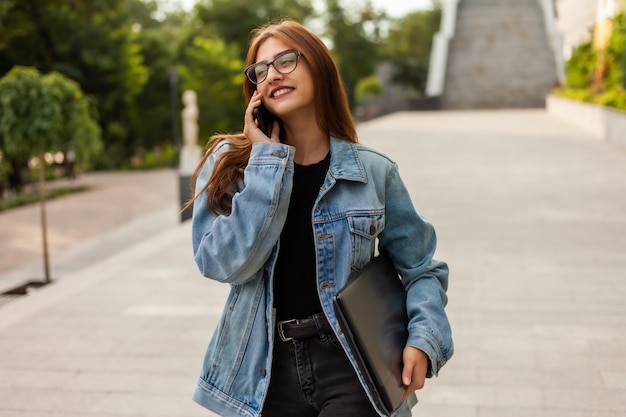 Tutti in affari. giovane donna alla moda in una giacca di jeans e occhiali in possesso di laptop e parlare al telefono mentre si cammina sulla città.