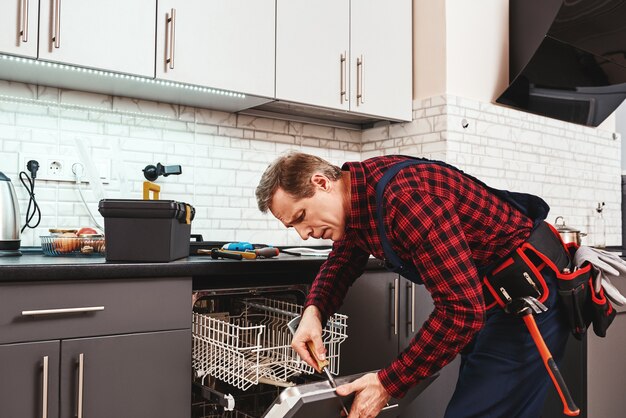 Tutti gli strumenti sono inclusi. tecnico maschio in piedi vicino alla lavastoviglie con cacciavite in cucina con strumenti