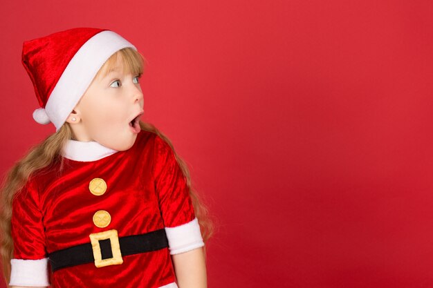 写真 私にとってこれらすべて！クリスマスの帽子とサンタの衣装を着て目をそらしている小さなかわいい女の子のプロフィールは、彼女の口を開いたコピースペースにショックを受けました