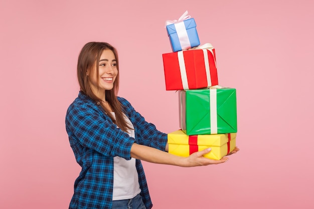 Alle cadeautjes voor jou Zijaanzicht van genereus aardig meisje dat veel geschenkdozen geeft en glimlacht feliciteren met kerstvakantie verjaardagsviering binnen studio opname geïsoleerd op roze achtergrond