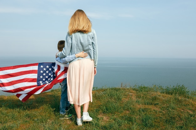 Alleenstaande moeder met zoon op onafhankelijkheidsdag van de VS Vrouw en haar kind lopen met de vlag van de Verenigde Staten aan de oceaan kust