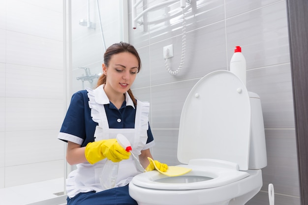 Alleenstaande vrouwelijke meid met vrolijke uitdrukking geknield op het toilet terwijl ze de stoel schoonmaakt met spuitfles en gele doek in rubberen gehandschoende handen