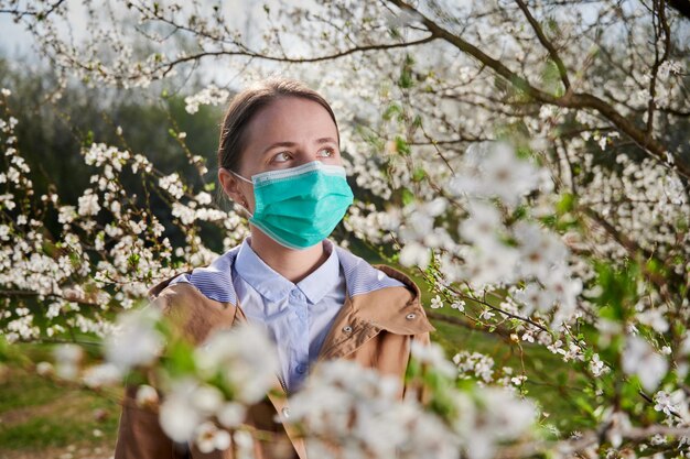 Allergische vrouw die lijdt aan seizoensgebonden allergie in de lente poserend in bloeiende tuin in de lente met medisch masker tussen bloeiende bomen Lente allergie concept