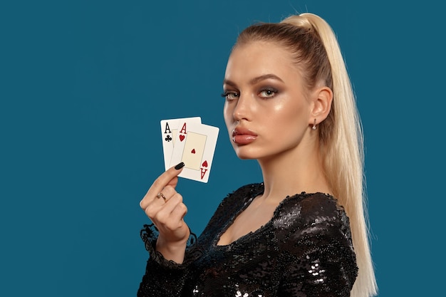 Alluring blonde model with ponytail, in jewelry and black sequin dress. She is showing two playing cards while posing sideways against blue background. Gambling entertainment, poker, casino. Close-up