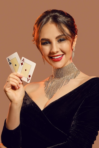 Alluring brunette female with bare shoulders, bright make-up, in black dress and brilliant jewelry. She is smiling, showing two aces, posing on brown background. Poker, casino. Close-up, copy space