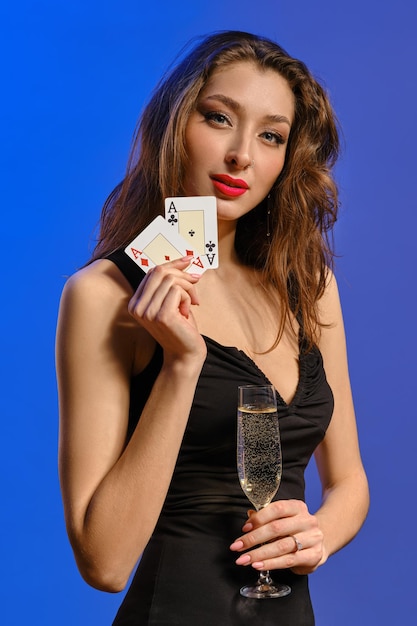 Alluring brunette lady with bright make-up and earring in nose,\
in black dress. she is holding a glass of champagne and two aces,\
smiling, posing on blue background. poker, casino. close-up
