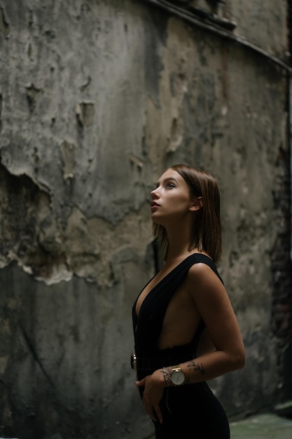 Очаровательная симпатичная девушка в черном эротическом топе стоит возле старой стены.