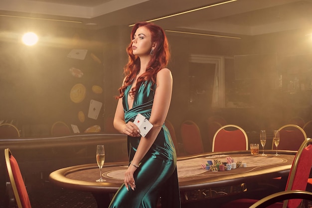 Alluring redheaded woman in a long blue satin dress is posing\
sideways with two aces in her hand against a poker table in luxury\
casino. passion, cards, chips, alcohol, win, gambling - it is as\
female