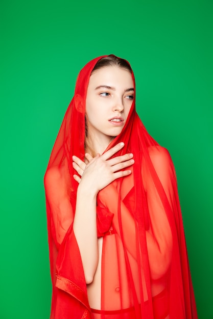 Alluring young slender fashionable female model in bright red underwear and headscarf standing with eyes closed against green background