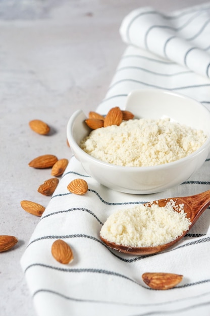 Almond flour in a bowl and spoon with scattered nuts on a white concrete background