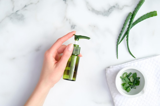 Photo aloe vera gel green plastic bottle in female hand on marble background with aloe plant stems natural organic lotion for hand and face skin treatment top view flat lay