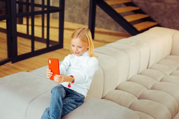 Alone at home. Charming kid sitting on sofa and looking at screen of her gadget