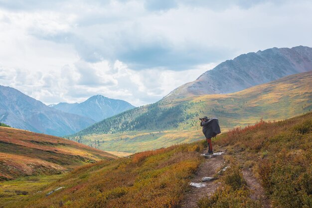 Alone traveler in raincoat with large backpack photographs nature among fading autumn vegetations on hiking trail through mountain pass Backpacker with photo camera shoots autumn mountain landscape