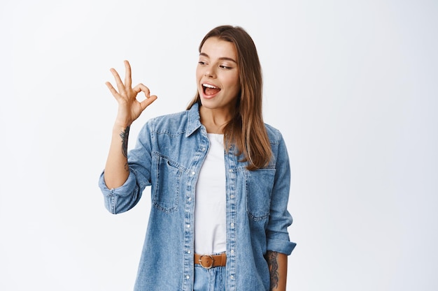 Bene. sorridente ragazza carina che mostra un segno ok e dice di sì, approva qualcosa di buono, piace e concorda, in piedi soddisfatta sul bianco