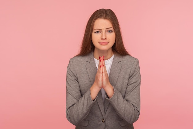 Alsjeblieft, ik heb hulp nodig. Portret van een smekende jonge vrouw in een pak biddend, smekend met ondeugende en smekende grimas, gebaar van verontschuldiging. studio-opname geïsoleerd op roze achtergrond