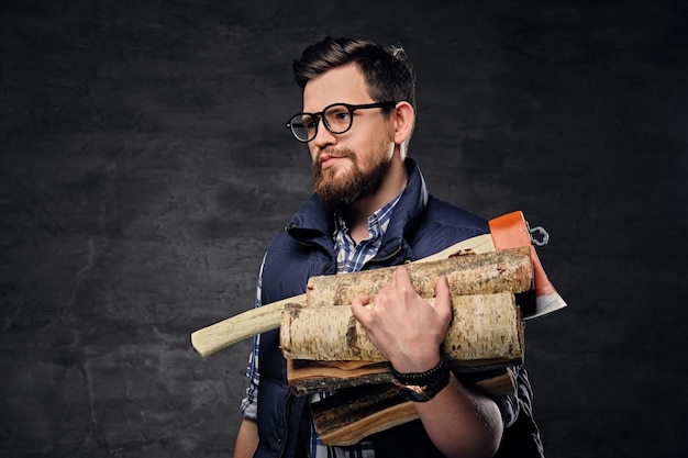 Alternative bearded male in eyeglasses dressed in fleece shirt holds firewoods and axe over grey background.