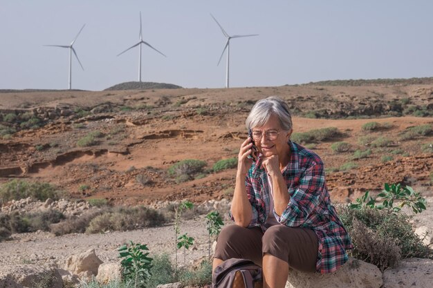 代替エネルギー風力発電所電話を使用しながら風力タービンの田園地帯に座っている年配の女性再生可能エネルギーの概念自然の愛クリーングリーン電力未来