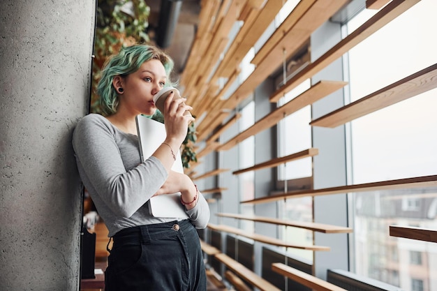 Foto ragazza alternativa in abiti casuali e con i capelli verdi in piedi con una tazza di bevanda in mano