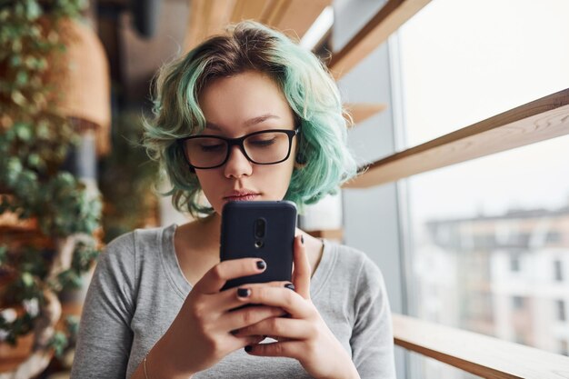 Alternative girl in glasses and with green hair sitting indoors at daytime with phone in hands