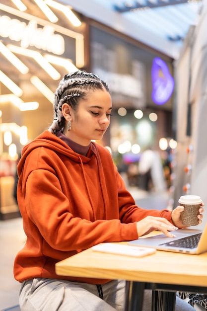 Alternative girl with white braids with a computer in a shopping center, looking at the camera with a hot coffee in her hands