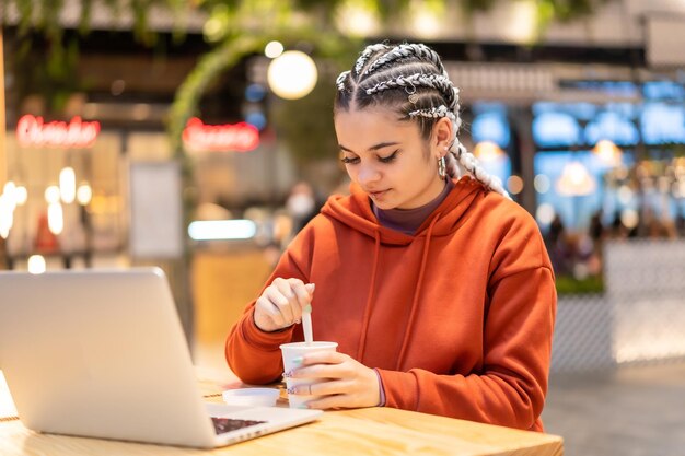 Alternative girl with white braids with a computer in a shopping center, making a video call with a hot coffee in her hands