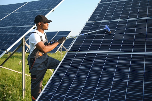 ユニフォームを着た代替発電所の労働者がモップで太陽電池パネルを掃除しているハンサムなアフリカ系アメリカ人が機器の世話をしている