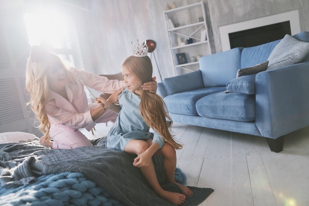 Altijd gezellig samen! Mooie jonge moeder die naar haar schattige dochter kijkt en glimlacht terwijl ze thuis op bed zit