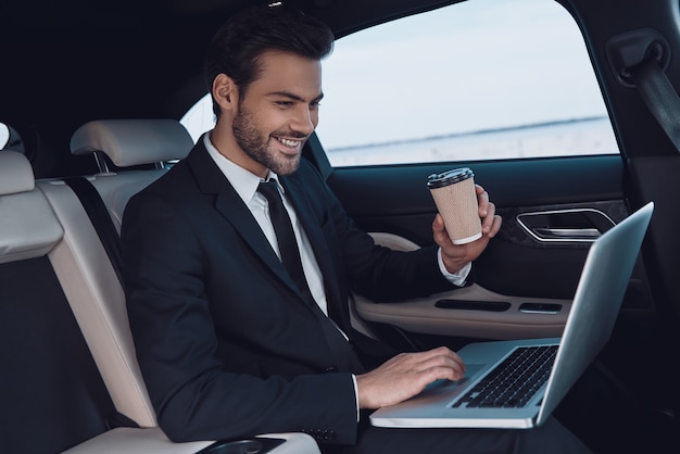 Altijd in contact. Knappe jongeman in volledig pak aan het werk met laptop terwijl hij in de auto zit