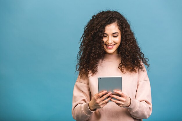 Altijd in contact! mooi krullend jong meisje glimlachend met behulp van tablet geïsoleerd op blauwe achtergrond.