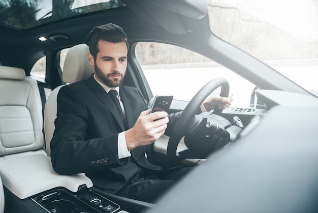 Always available. Handsome young businessman looking on his smart phone while sitting on the front seat