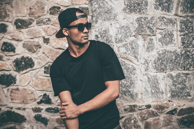 Always cool and trendy. Handsome young African man in cap and sunglasses standing against the stoned wall and looking away