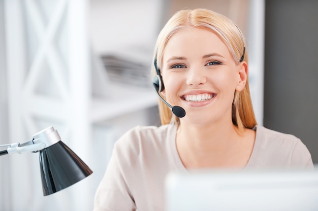 Always ready to help you. Happy young woman in headset looking at camera and smiling while sitting at her working place