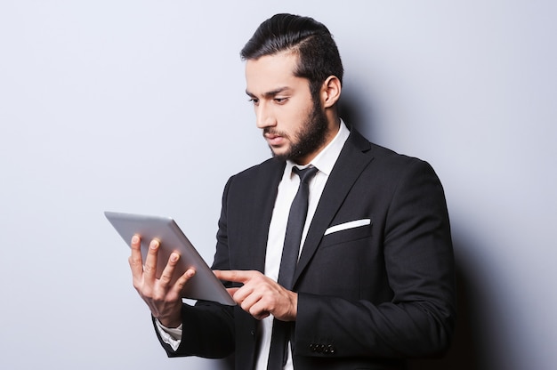 Always serious while working. Confident young man in formalwear working on digital tablet while standing against grey background