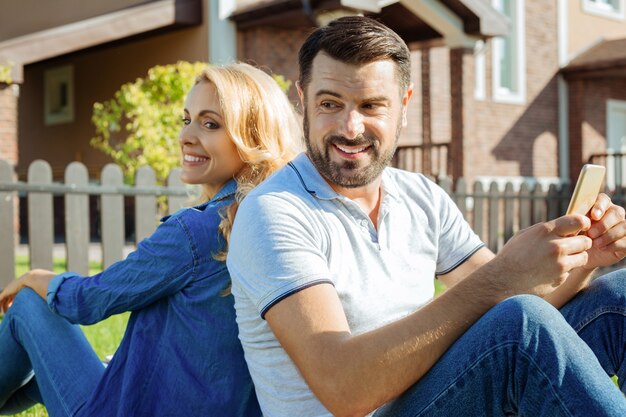 Always together. Lovable middle-aged couple sitting back-to-back on the grass, turning their heads to each other and smiling while the man holding a phone