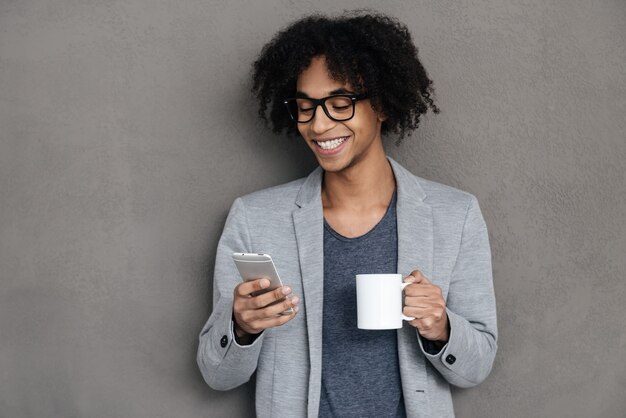 Всегда на связи. Веселый молодой африканский мужчина держит смартфон и смотрит на него с улыбкой, стоя на сером фоне с чашкой кофе
