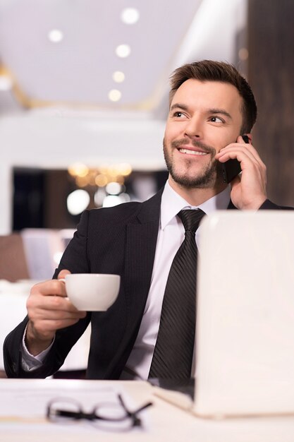 Always in touch. Cheerful young man in formalwear talking on the phone and drinking coffee while sitting at the restaurant