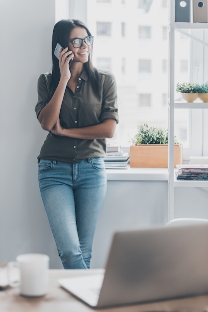 Always in touch. Confident young woman in smart casual wear talking on the mobile phone and smiling while standing near her working place in office