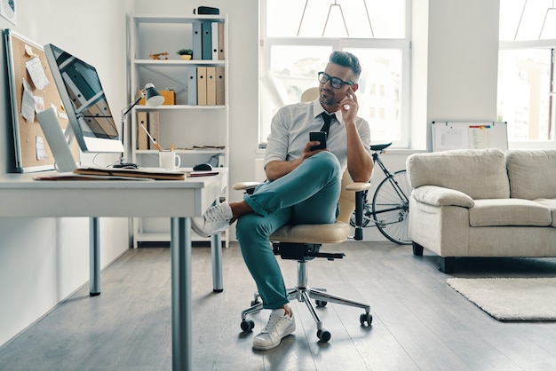 Sempre in contatto. bel giovane in camicia e cravatta che usa lo smartphone e sorride mentre è seduto in ufficio