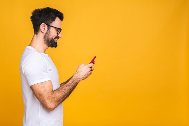 Always in touch. Happy young man in glasses typing sms isolated on yellow background, smiling and looking at camera.