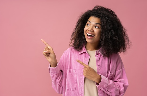 Amazed African American woman wearing pointing finger on copy space isolated on pink background