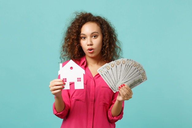 Amazed african girl in casual clothes holding paper house, fan of money in dollar banknotes, cash money isolated on blue turquoise background. People emotions, lifestyle concept. Mock up copy space.