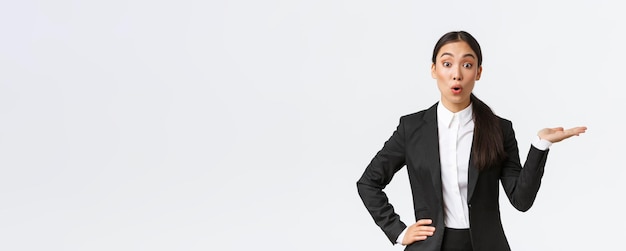 Amazed asian businesswoman in black suit pointing hand right and look at camera interested discussing announcement showing banner standing white background intrigued Copy space