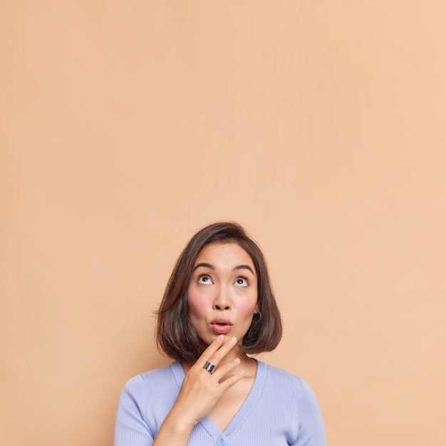 Amazed Asian woman with dark hair focused above reacts on something shocking keeps hand on chin wears blue jumper poses against beige wall blank copy space overhead for your promotion