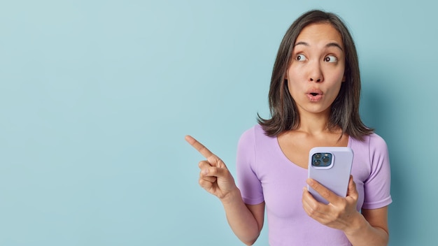 Amazed asian woman with dark hair points aside shows\
advertisement uses mobile phone enjoys online communication dressed\
in casual purple t shirt isolated over blue background technology\
and reaction