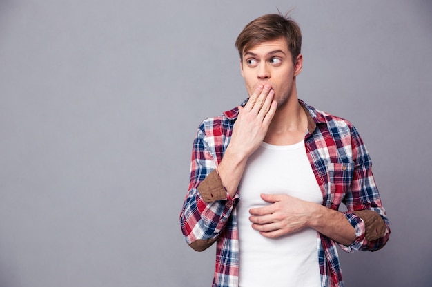 Amazed astonished young man in plaid shirt covered mouth by hand and feeling afraid over grey wall