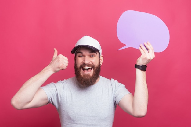 Amazed bearded man showing thumbs up and holding speech bubble