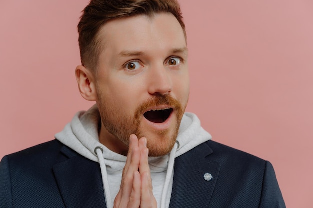 Photo amazed bearded man in sweatshirt and black jacket mouth open palms pressed together disbelief isolated on pink background